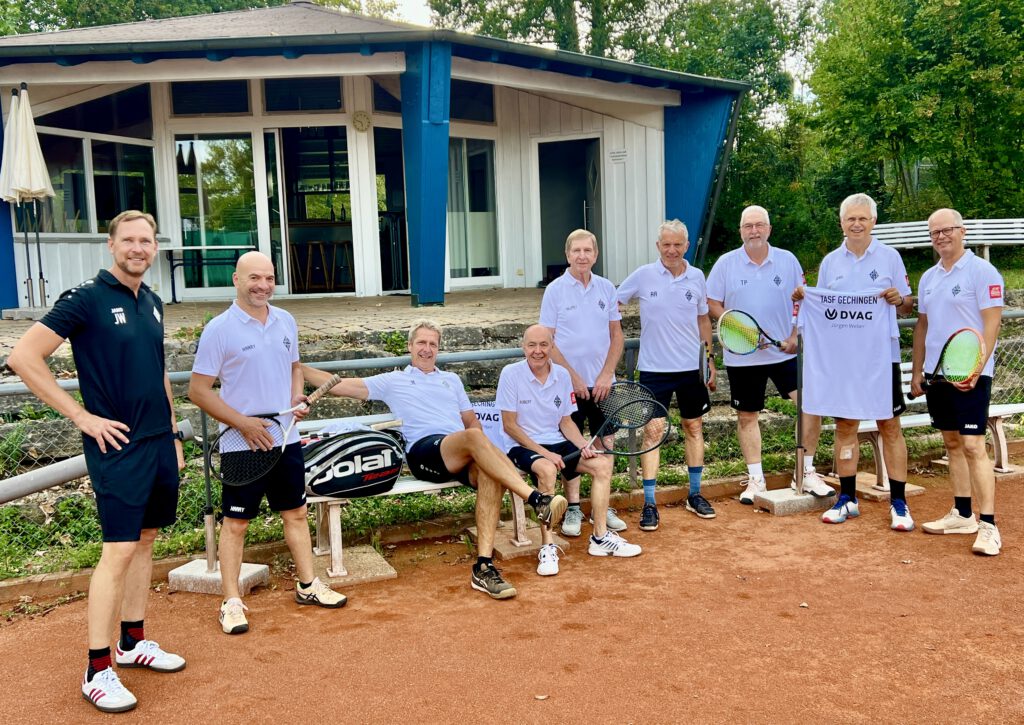 Mitglieder der Herren-50-Tennismannschaft der TASF Gechingen und Sponsor Jürgen Weber posieren in weißer Teambekleidung vor dem frisch sanierten blau-weißen Pavillon des Vereins. Ein Spieler sitzt auf einer Holzbank mit Tennistasche, die anderen stehen daneben, teils mit Schlägern in der Hand; einer präsentiert ein weißes Sponsoren-Shirt mit dem Aufdruck „TASF Gechingen" und dem DVAG-Logo.